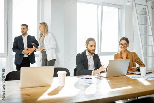 Group of a young office employees dressed casually in the suits having some office work at the large meeting table in the bright sunny room