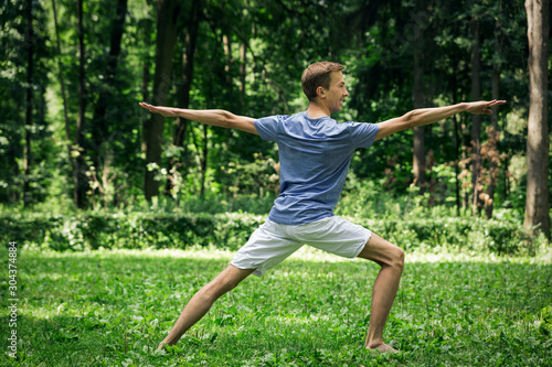 Young attractive man in a gray T-shirt and sweatpants doing yoga warrior pose in the park. He spread his legs wide, arms outstretched to the sides, looking to the side.