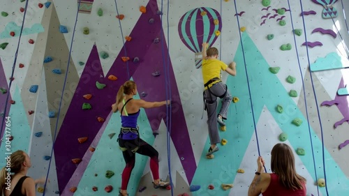 Wide tracking shot of teenage boy and young woman in safety gear, with ropes climbing up artificial wall at indoor gym, and their companions standing on ground and belaying photo