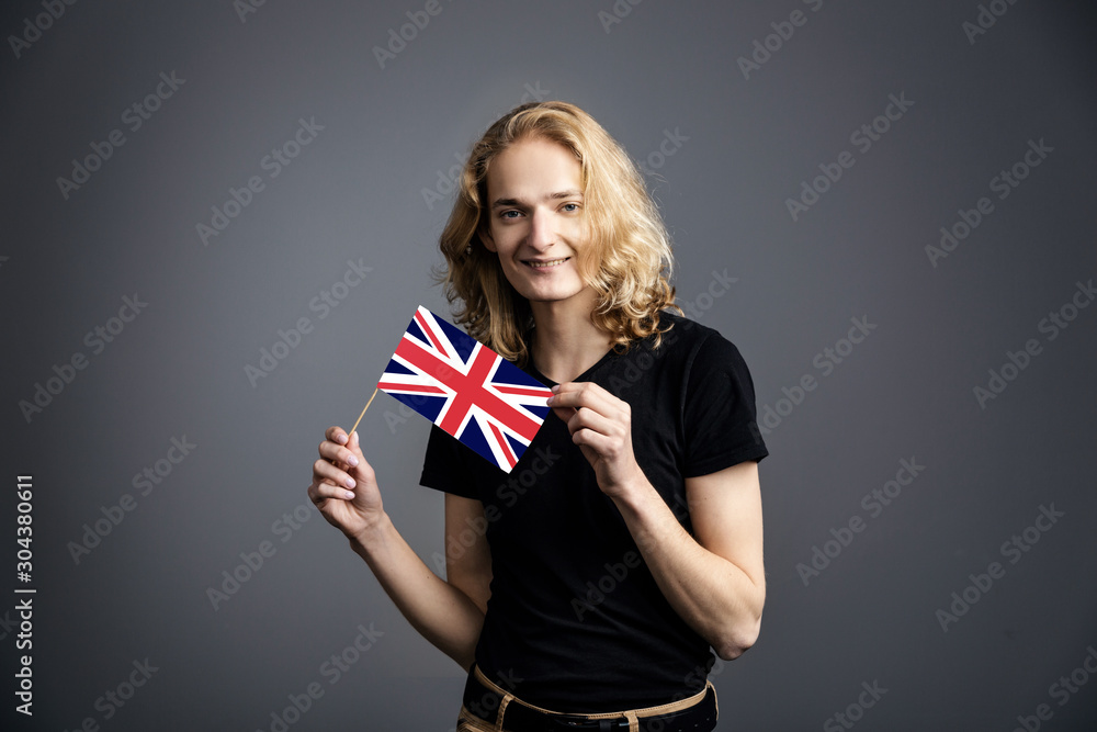 Fototapeta premium A young guy with long blonde hair in a nuclear t-shirt smiles and waves the UK flag on a gray background.