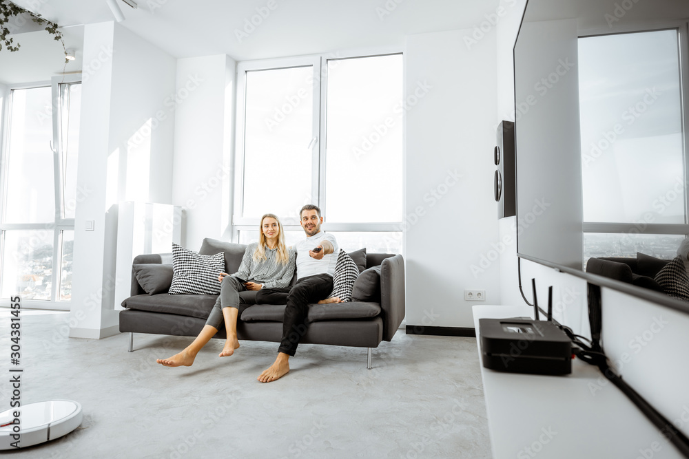 Young couple relaxing on the comfortable couch, watching television in the bright living room of the modern apartment Young couple relaxing on the comfortable couch, watching television in the bright living room of the modern apartment