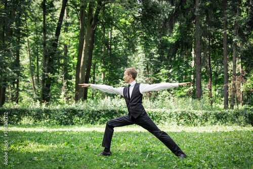 Attractive young man in a suit does yoga (warrior pose) in a park.