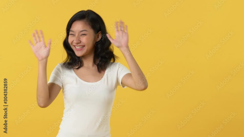 Cute happy smiling Asian woman in white t-shirt waving and dancing left and right against yellow background Cute happy smiling Asian woman in white t-shirt waving and dancing left and right against yellow background