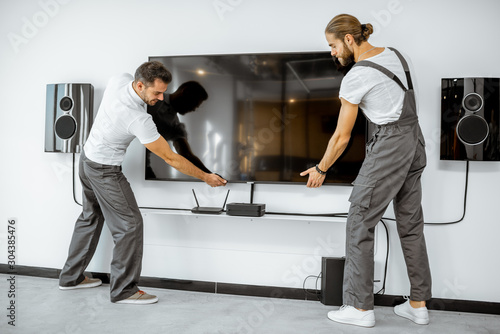 Two professional workmen in workwear installing a large TV monitor and audio system in the white living room. Home appliances installation concept