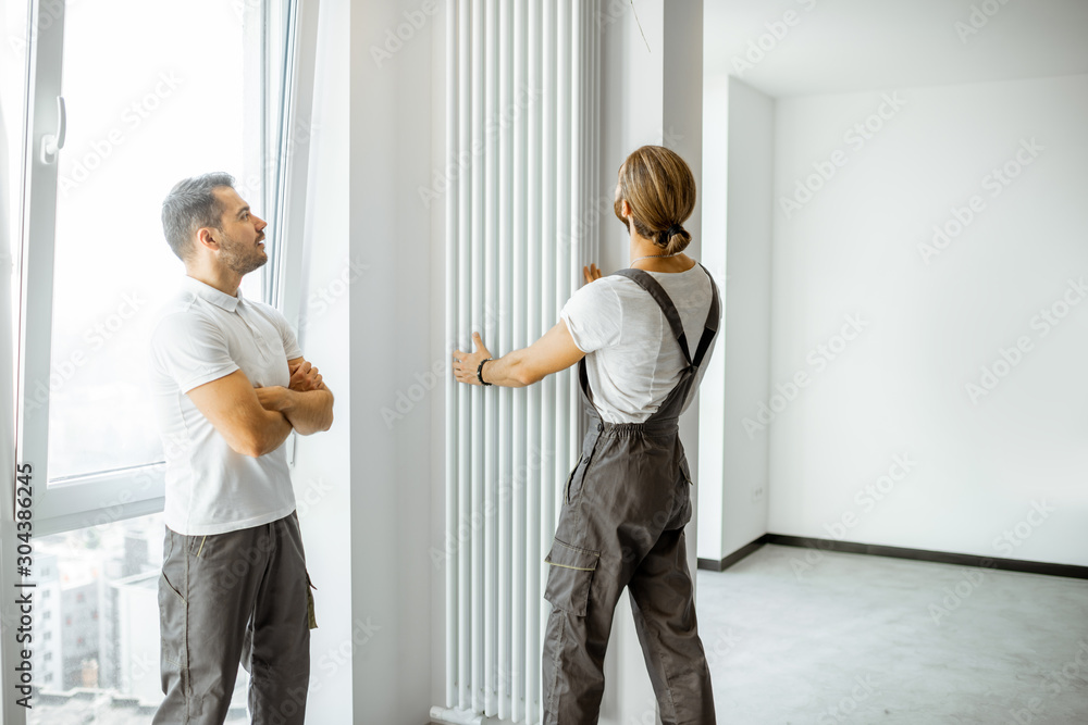 Fototapeta premium Two professional workmen in workwear installing decorative radiator in the white living room, finishing the repair in the apartment