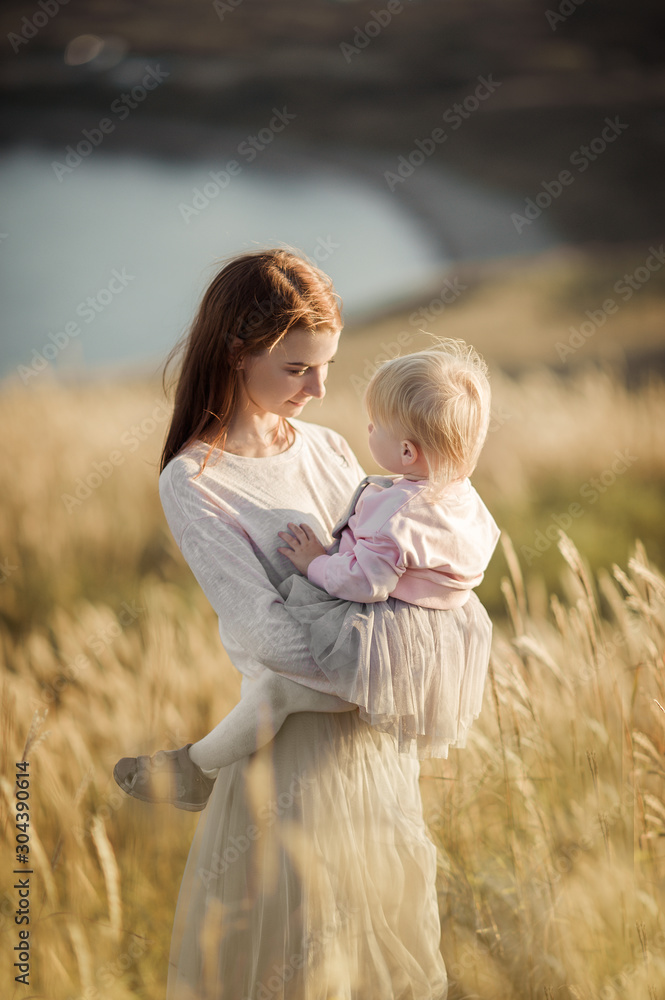 mother with red hair in pastel clothes holds a blonde daughter in her arms in a lush tulle skirt in a wheat field, in a sunny warm sunset light mother with red hair in pastel clothes holds a blonde daughter in her arms in a lush tulle skirt in a wheat field, in a sunny warm sunset light