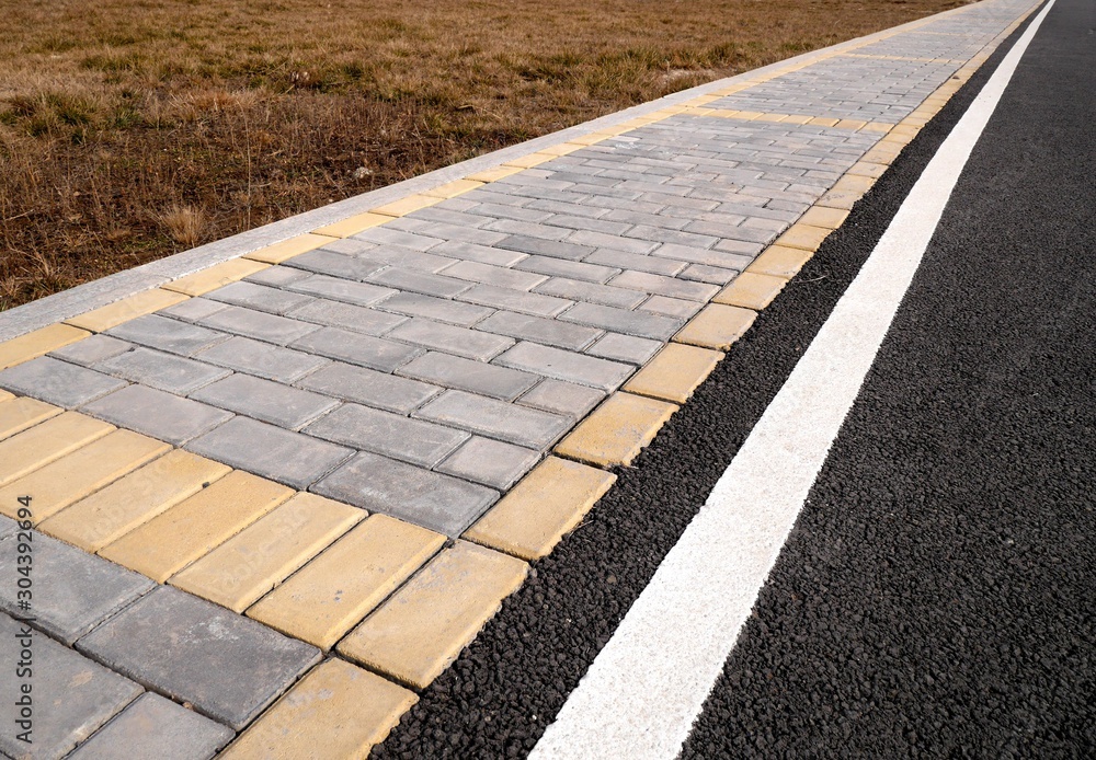 Top view photo of a fragment of a sidewalk paved with grey and yellow tiles and a bicycle lane with rubber coating, with dry grass .