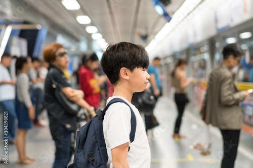 Smart looking, confident Asian preteen boy wear white t-shirt, backpack, wait for underground train in crowded platform in the morning, Routine commute of school student, city lifestyle, life skills. 