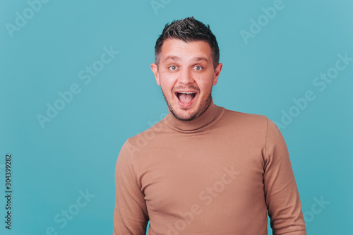 Portrait of happy cheerful playful fashionable man with appealing smile in beige sweater look at cam on blue background. Positive funny emotions, white teeth, laugh happily. Lifestyle concept