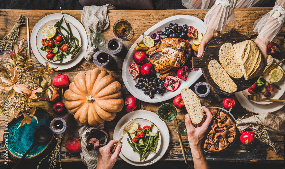 Thanksgiving party table setting. Flat-lay of whole roasted chicken, vegetables, fig pie, fruit, pumpkin, candles, tableware and peoples hands with food over rustic wooden table background, top view