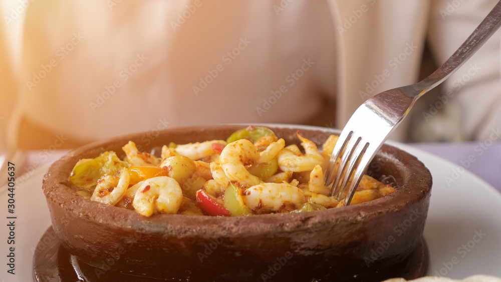 Good served food in restaurant. Woman is eating frying hot shrimps with vegetables served in clay bowl, closeup dish. Takes shrimp out of the boiling oil with a fork. Good served food in restaurant. Woman is eating frying hot shrimps with vegetables served in clay bowl, closeup dish. Takes shrimp out of the boiling oil with a fork.