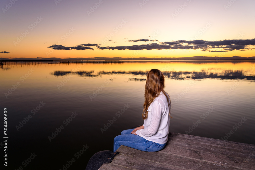 Long-haired young woman relaxing admiring the golden sunset on a beautiful pier by a natural lake.