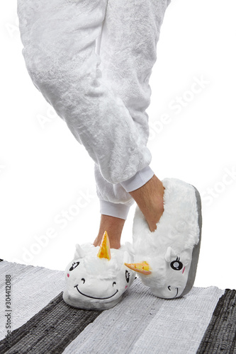 Close-up shot of male legs in white velour trousers and plush house slippers made in the form of white smiling unicorn. The man is standing on the striped gray and white carpet.