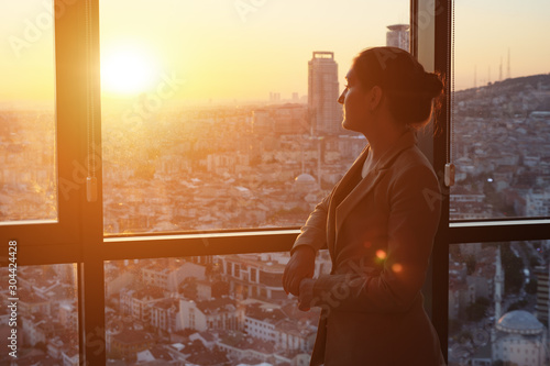 фотографія Young successful businesswoman in office suit standing near the window with panoramic city view in her office in evening, sunlight