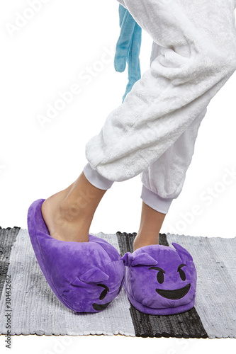 Close-up shot of female legs in white velour trousers and purple plush house slippers made in the form of the smiling devil emoji. The girl is standing on the striped gray and white carpet.