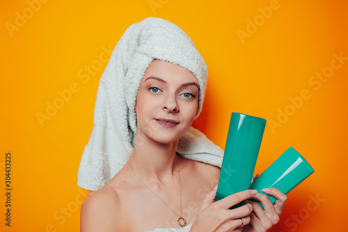 Young woman in white towel on head on the orange background holds shampoo bottle without logo. Woman in bath with clean and soft skin.