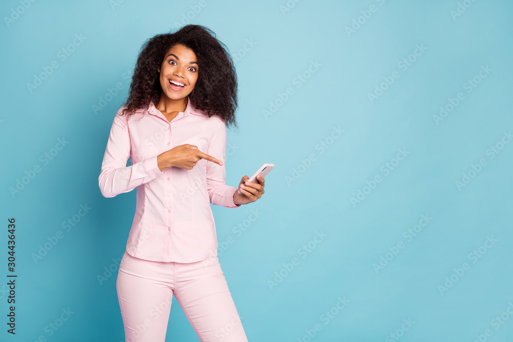 Photo of amazing wavy dark skin lady holding telephone hands indicating finger on screen sale smart phone price wear pink shirt pants isolated blue color background Photo of amazing wavy dark skin lady holding telephone hands indicating finger on screen sale smart phone price wear pink shirt pants isolated blue color background