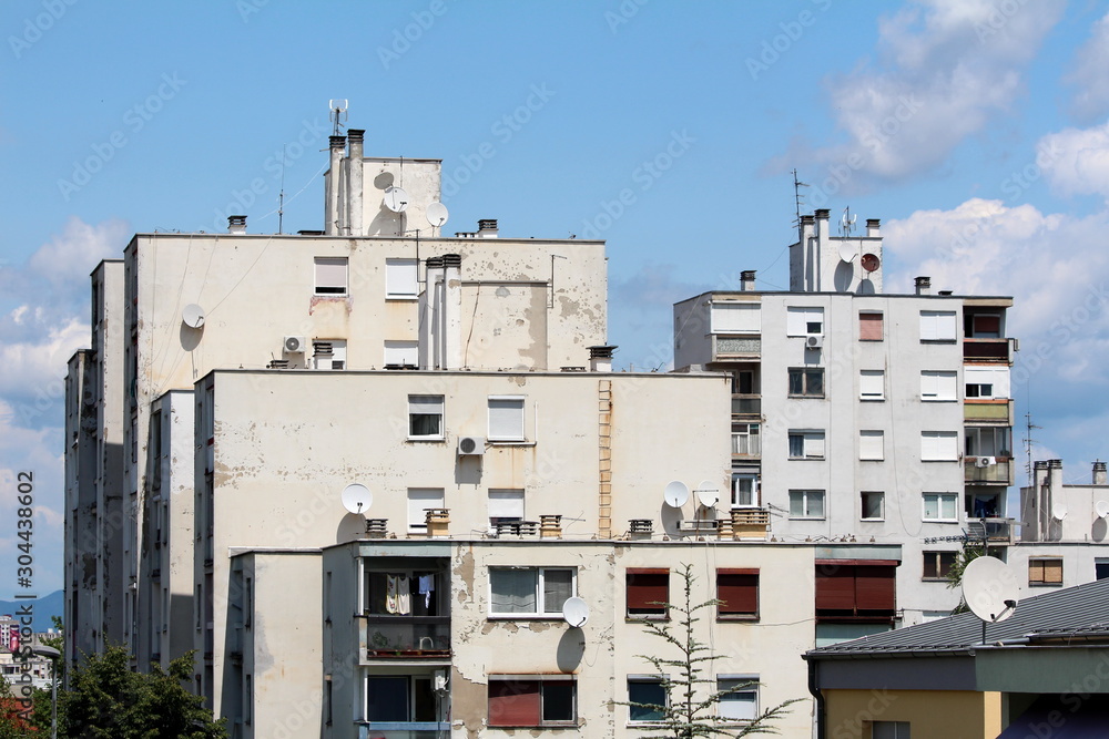 White dilapidated apartment buildings in old part of town with multiple satellite dishes and chimneys in various sizes on top on cloudy blue sky background of warm sunny summer day