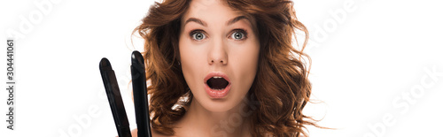 panoramic shot of surprised curly girl holding hair iron isolated on white