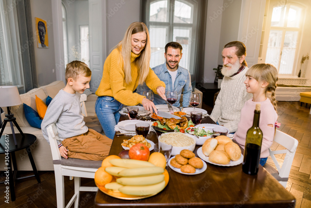 Family of five people, grandfather, parents and children sitting at the table and going to eat roasted turkey, while happy mother is cutting turkey. Thanksgiving family dinner concept