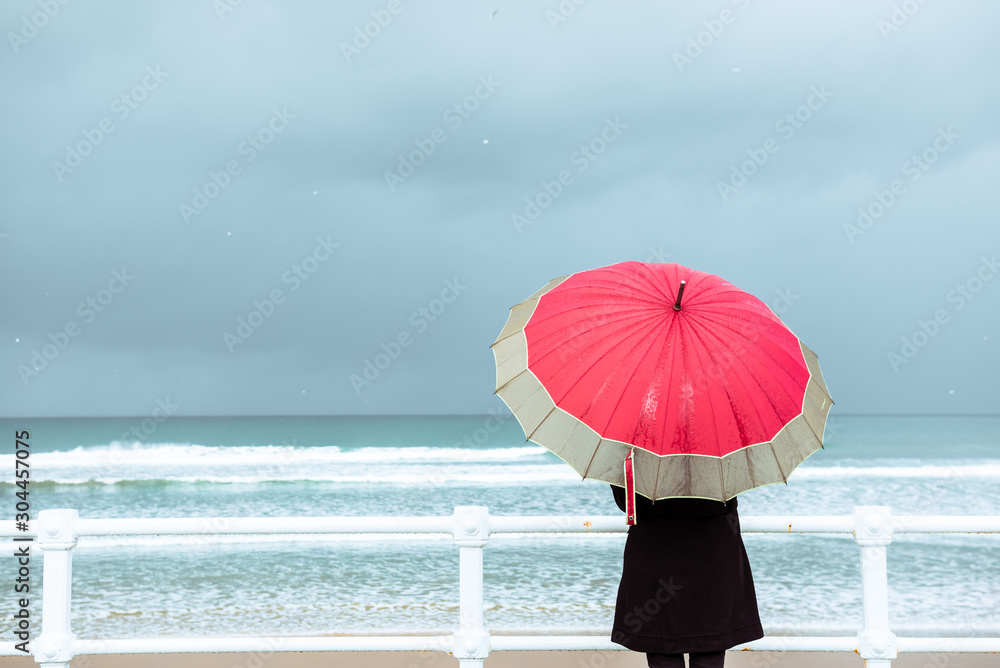 A woman watches the sea on a rainy day. She is facing away from the camera, facing the sea and with a red umbrella. Vertical stock image.