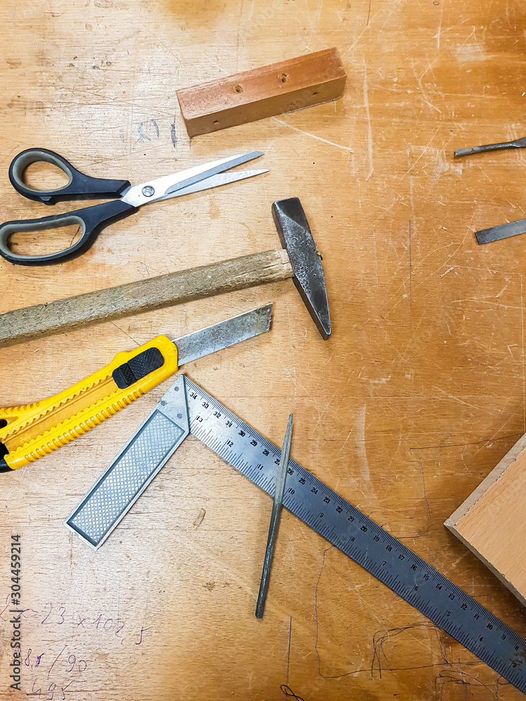 Various workshop tools: box cutter knife, scissors, hammer and a metal ruler on a traditional wooden work table. Crafting carpentry work space crafts creative background concept, top view Various workshop tools: box cutter knife, scissors, hammer and a metal ruler on a traditional wooden work table. Crafting carpentry work space crafts creative background concept, top view