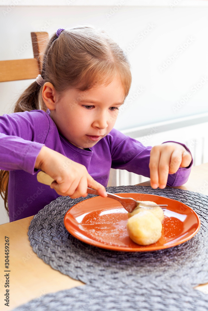 Primary school young girl sitting on a chair near a table, eating potato dumplings all by herself, holding a fork in her hands cutting the food. Child using cutlery concept, finishing her meal Primary school young girl sitting on a chair near a table, eating potato dumplings all by herself, holding a fork in her hands cutting the food. Child using cutlery concept, finishing her meal