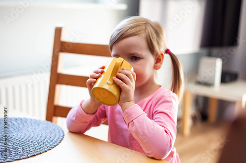 Young preschool girl with ponytails standing near a table, child drinking from a yellow cup / mug by herself, holding the beverage in two hands elbows on the table living room and tv in the background