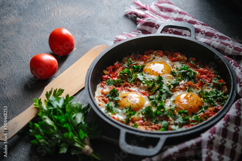 Traditional shakshuka with eggs, tomato, and parsley in a iron pan on a dark background