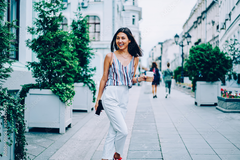 Half length portrait of cheerful woman enjoying city tour on holiday holding coffee to go, excited asian girl dressed in stylish apparel walking on street in downtown with takeaway happy about leisure Half length portrait of cheerful woman enjoying city tour on holiday holding coffee to go, excited asian girl dressed in stylish apparel walking on street in downtown with takeaway happy about leisure