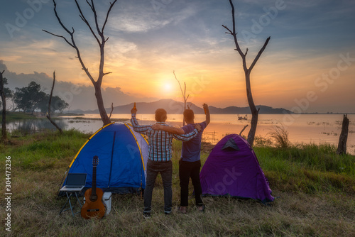 young friends camping by playing guitar and drinking beverage to happily near tent with lake background.
