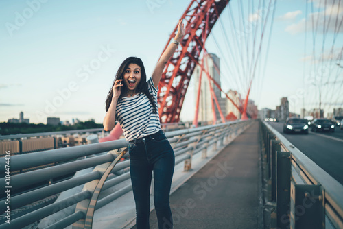 Half length portrait of wonder hipster girl with raised hand satisfied with received news during cellphone communication, excited Asian woman surprised with incoming international phone call