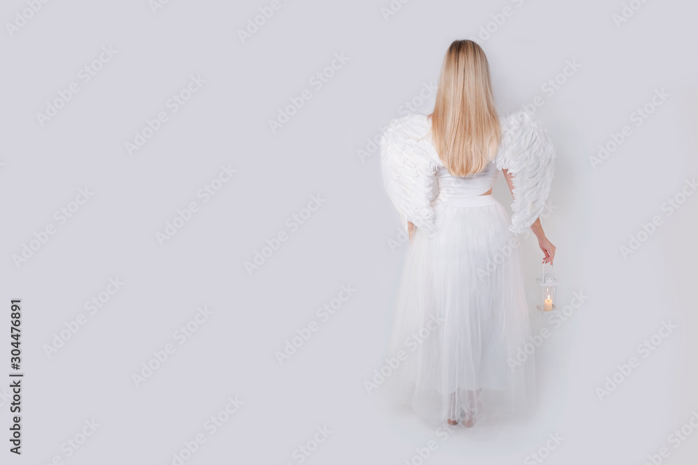 Girl blonde angel in white and with wings holds a candlestick in her hands and stands with her back. Isolated angel with wings. Girl blonde angel in white and with wings holds a candlestick in her hands and stands with her back. Isolated angel with wings.
