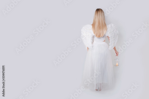 Girl blonde angel in white and with wings holds a candlestick in her hands and stands with her back. Isolated angel with wings.
