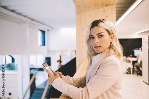 Portrait of confident female lawyer standing in modern workshop with smartphone device in hand and looking at camera, caucasian businesswoman using cellular phone during working time indoors