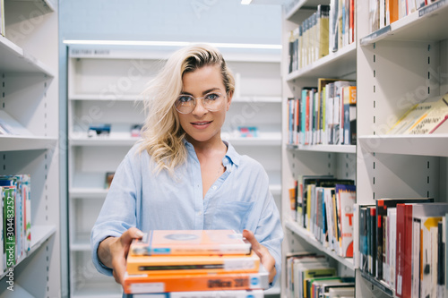 Half length portrait of young positive female librarian in trendy eyeglasses for vision correction holding books for education and offering them for reading, concept of knowledge and education