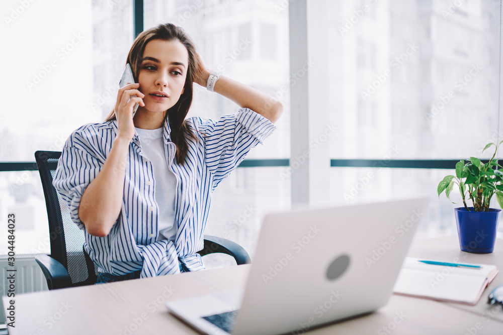 Pensive young woman calling to operator of customer support update software on laptop computer in office,serious female freelancer concentrated on telephone conversation about online business.