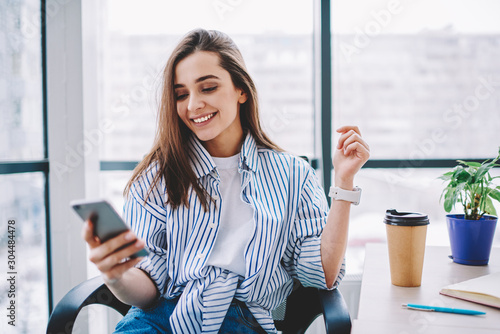 Cheerful young woman checking new feed and content in social networks via smartphone during coffee break in office happy cute female blogger satisfied with followers messages sitting at desktop.