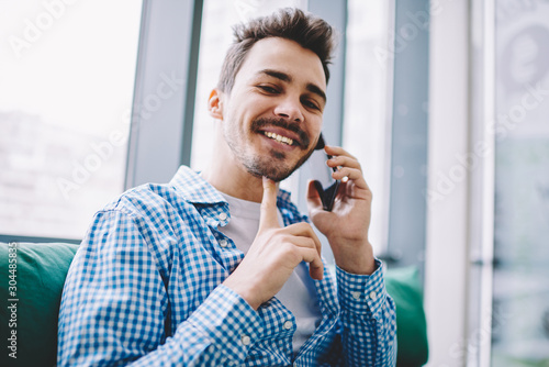 Portrait of cheerful hipster guy looking at camera while enjoying break time for making positive smartphone conversation with best friend using 4g wireless connection and mobile application indoors