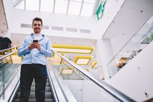 Half length portrait of cheerful man looking at camera while going down on modern escalator on publicity area holding smartphone for checking balance on credit card via application before shopping