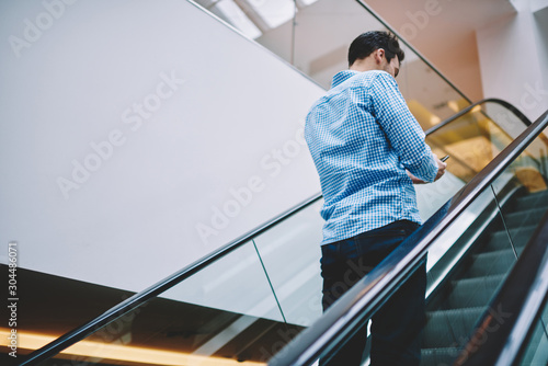 Back view of millennial hipster guy standing on escalator in shopping center and using public wireless connection for browsing internet, male blogger checking messages from followers via cellphone