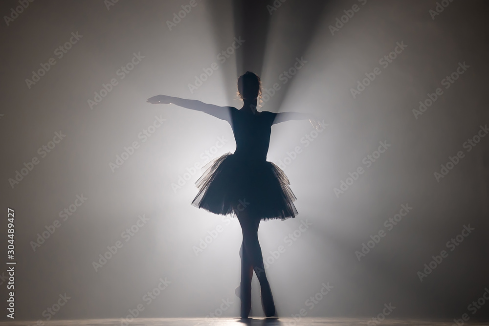 Fototapeta Profesjonalny balerina taniec balet w reflektorach dymu na dużej scenie. Piękna młoda dziewczyna ubrana w czarną sukienkę tutu na tle reflektorów. Czarny i biały.