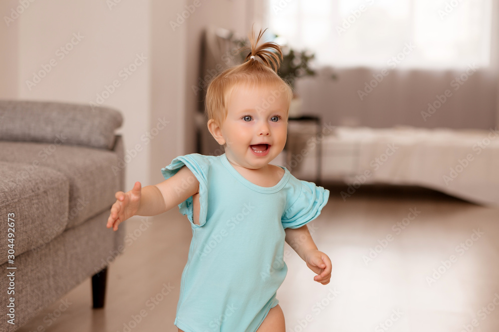 healthy baby girl in a blue bodysuit in a room next to a gray sofa is learning to walk. the comfort of the home. the child smiles healthy baby girl in a blue bodysuit in a room next to a gray sofa is learning to walk. the comfort of the home. the child smiles