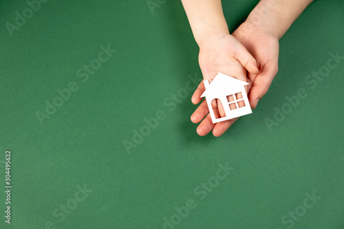 Top view of female and child hands holding white paper house on green background. Family home and real estate concept. Flat lay, copy space