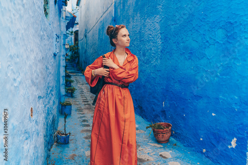 a tourist in a long bright orange dress with a backpack walks through the blue city of Morocco © nelen.ru