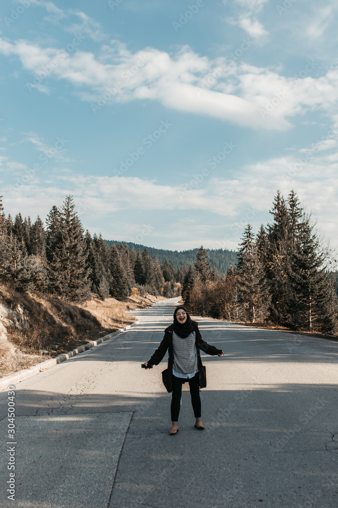 Woman with hijab goes along the line of road markings. She walks in the center of a quiet road in the mountain, surrounded by trees. Mountain Igman, Bosnia and Herzegovina. It is hipster travelling. Woman with hijab goes along the line of road markings. She walks in the center of a quiet road in the mountain, surrounded by trees. Mountain Igman, Bosnia and Herzegovina. It is hipster travelling.