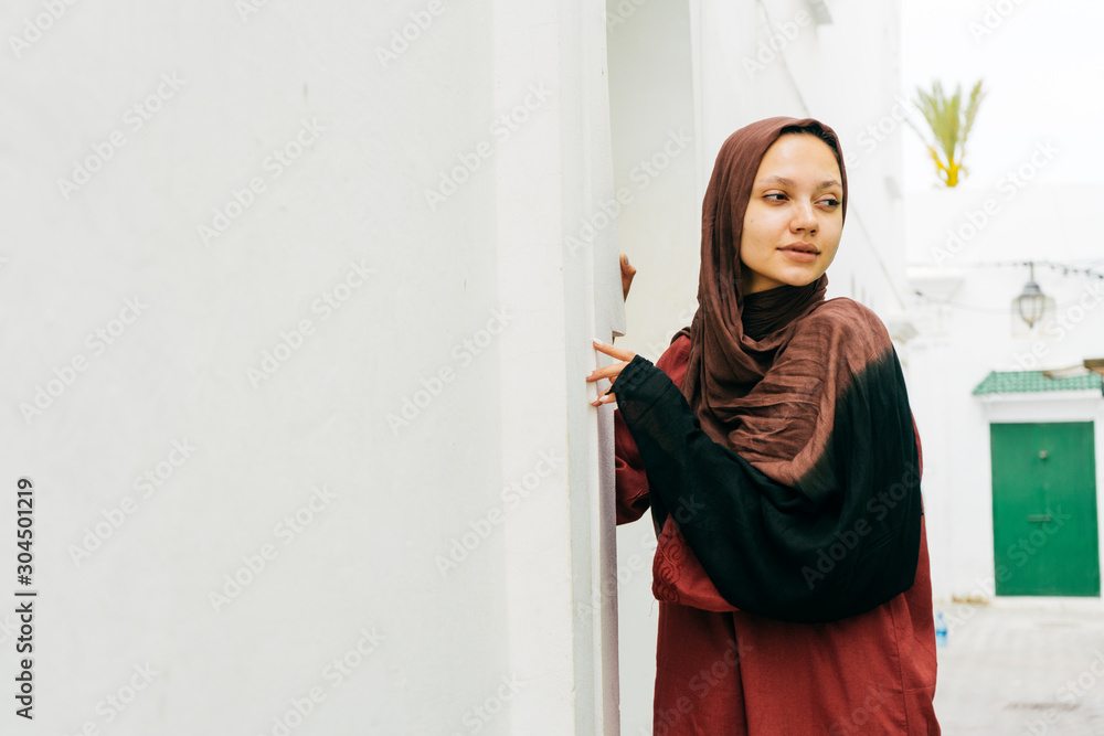 a muslim woman without makeup stands by a white-painted wall and looks timidly around a muslim woman without makeup stands by a white-painted wall and looks timidly around