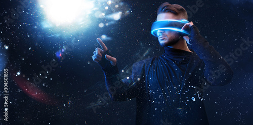 Beautiful young man using VR helmet while touching air in colorful neon lights. Guy in glasses of virtual reality with blue backlight over dark magic universe background. 