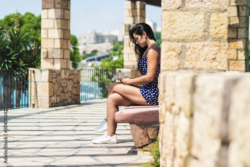 Beautiful hispanic woman in blue dress sitting on a bench while using a smartphone