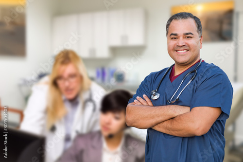 Handsome Hispanic Doctor or Nurse Standing in His Office with Staff Working Behind